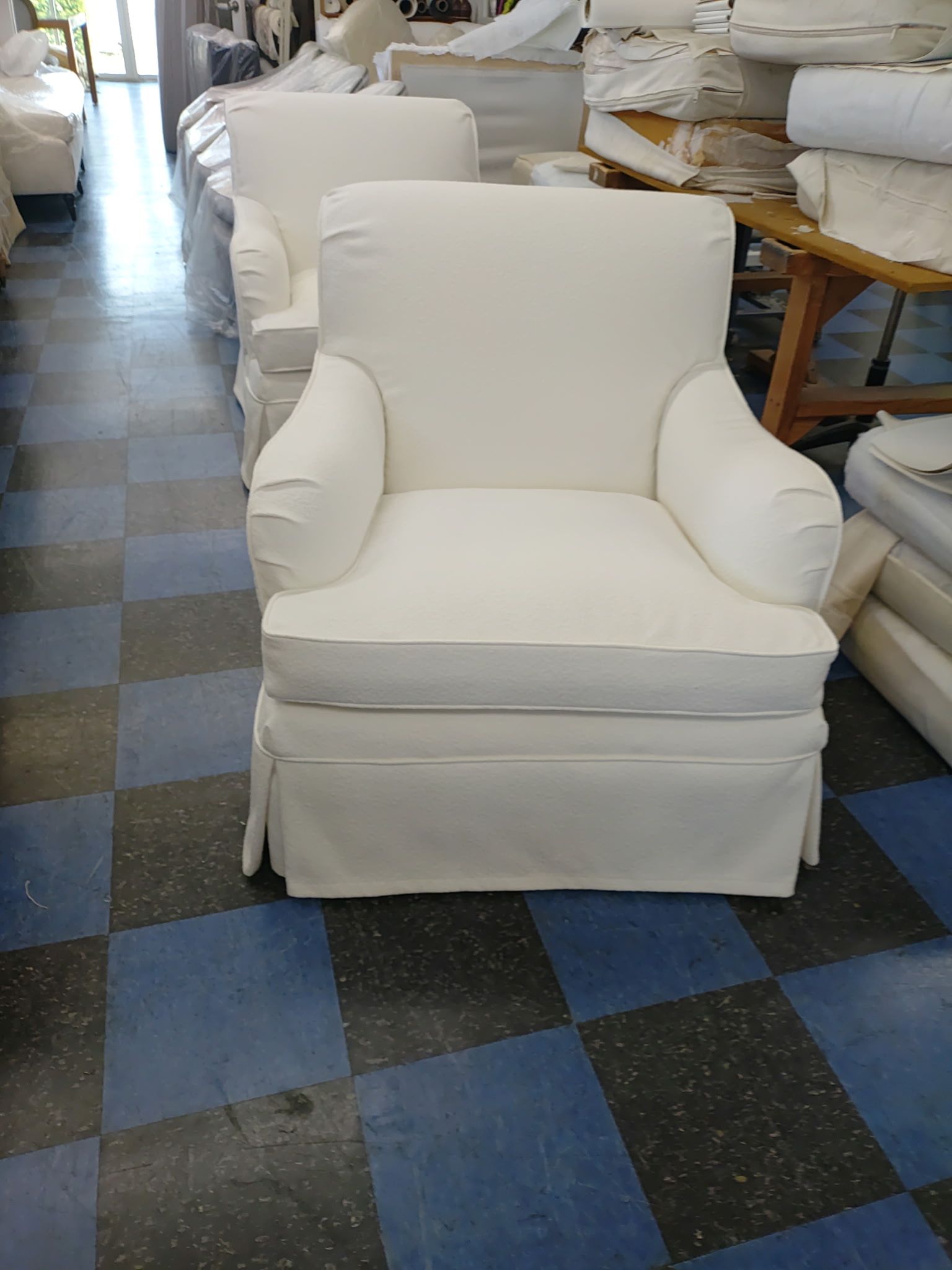 An off-white upholstered armchair with a skirted base, situated in a workshop with a checkered floor.
