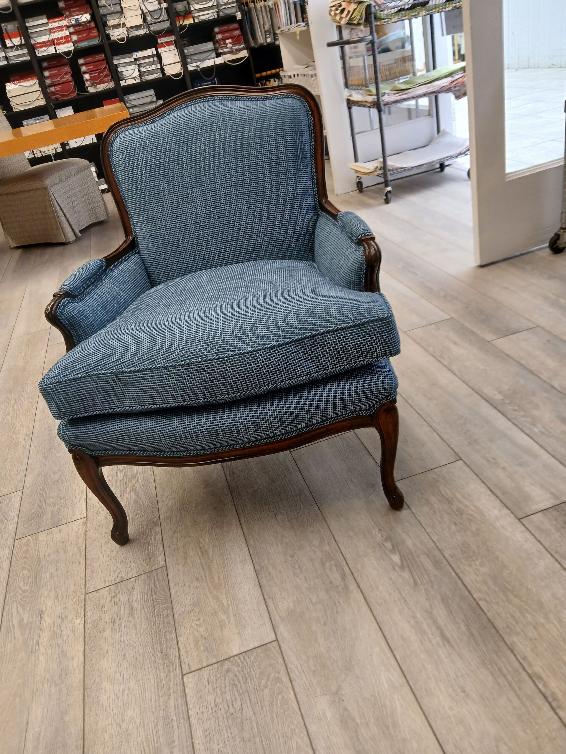 A blue patterned armchair with a dark wooden frame sits on a light-colored wooden floor in a shop.