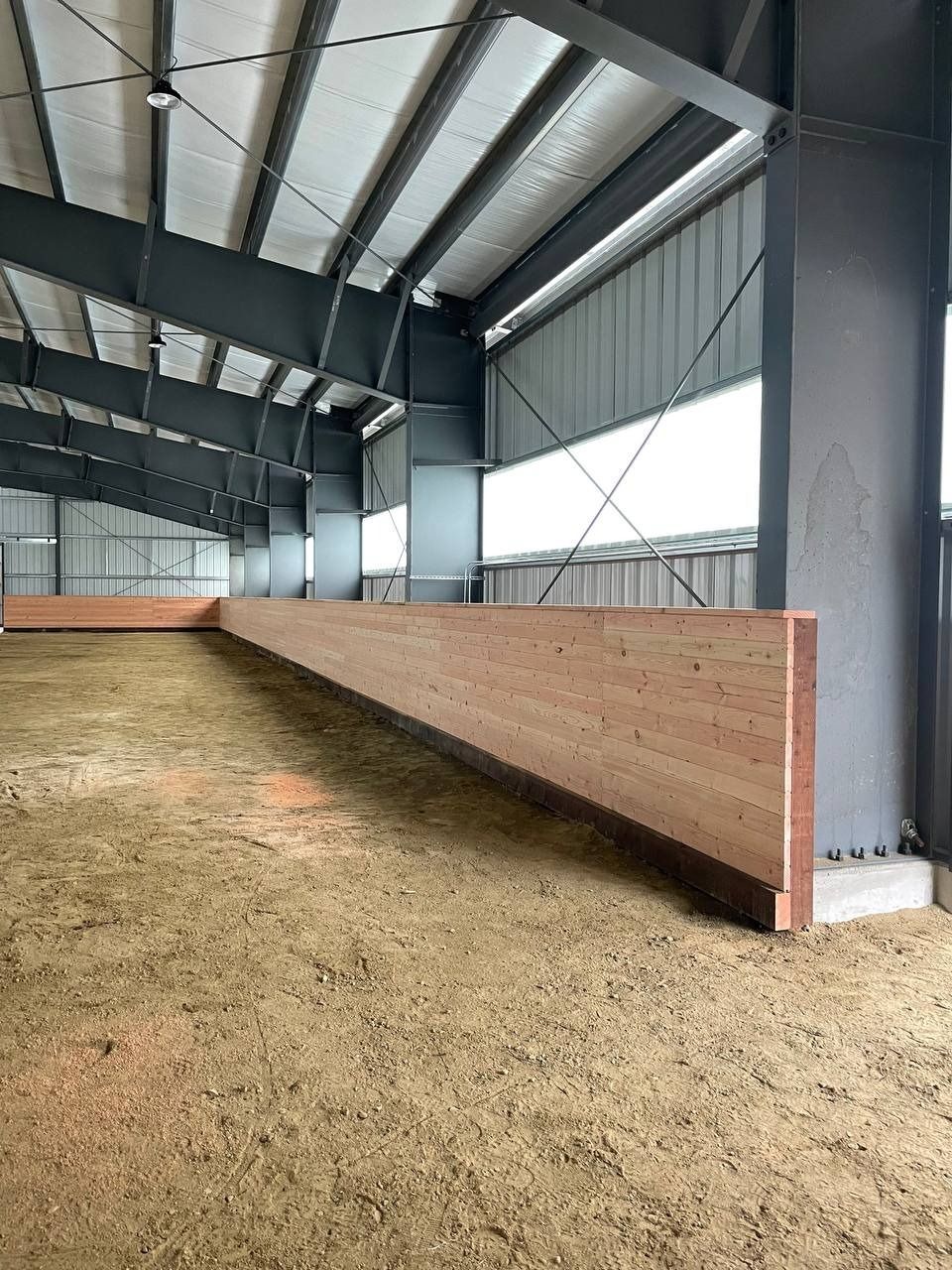 Indoor equestrian arena with sand footing, wooden kickboards, and steel supports.