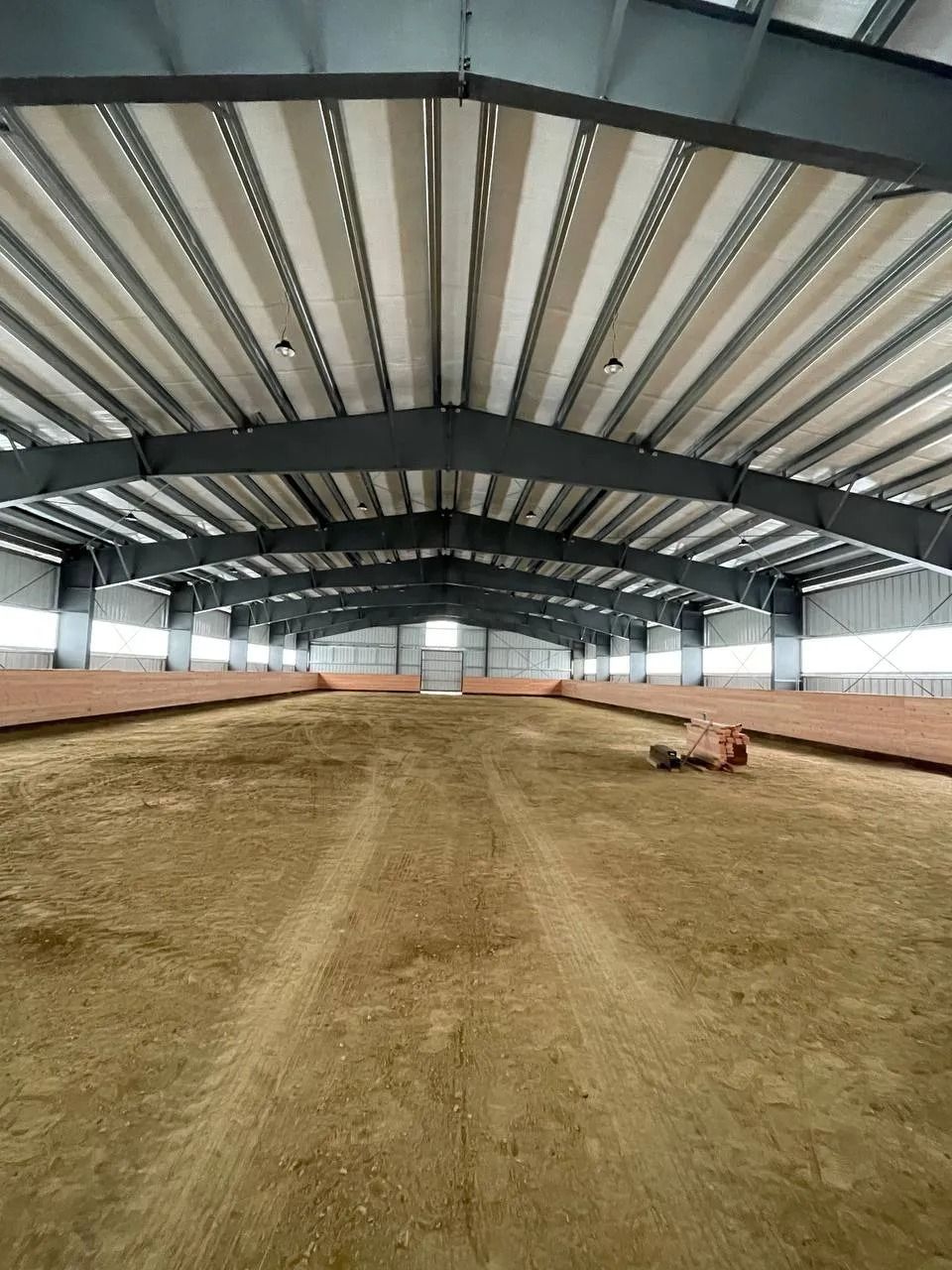 Indoor equestrian arena with dirt floor, metal supports, and bright ceiling.