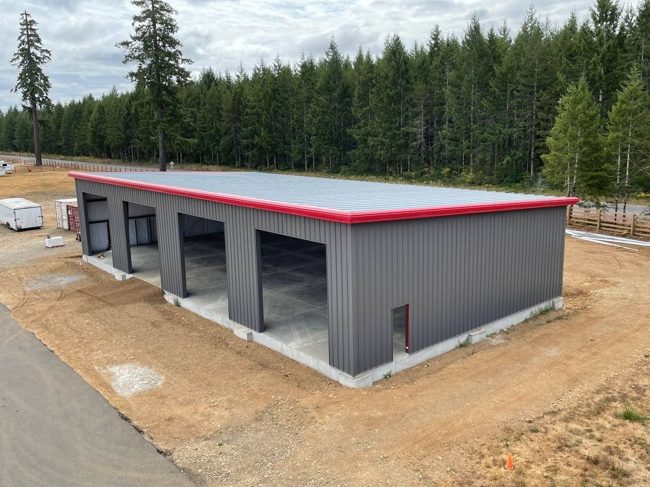Gray metal garage with three bays, red trim, and concrete foundation. Situated on gravel with trees in the background.