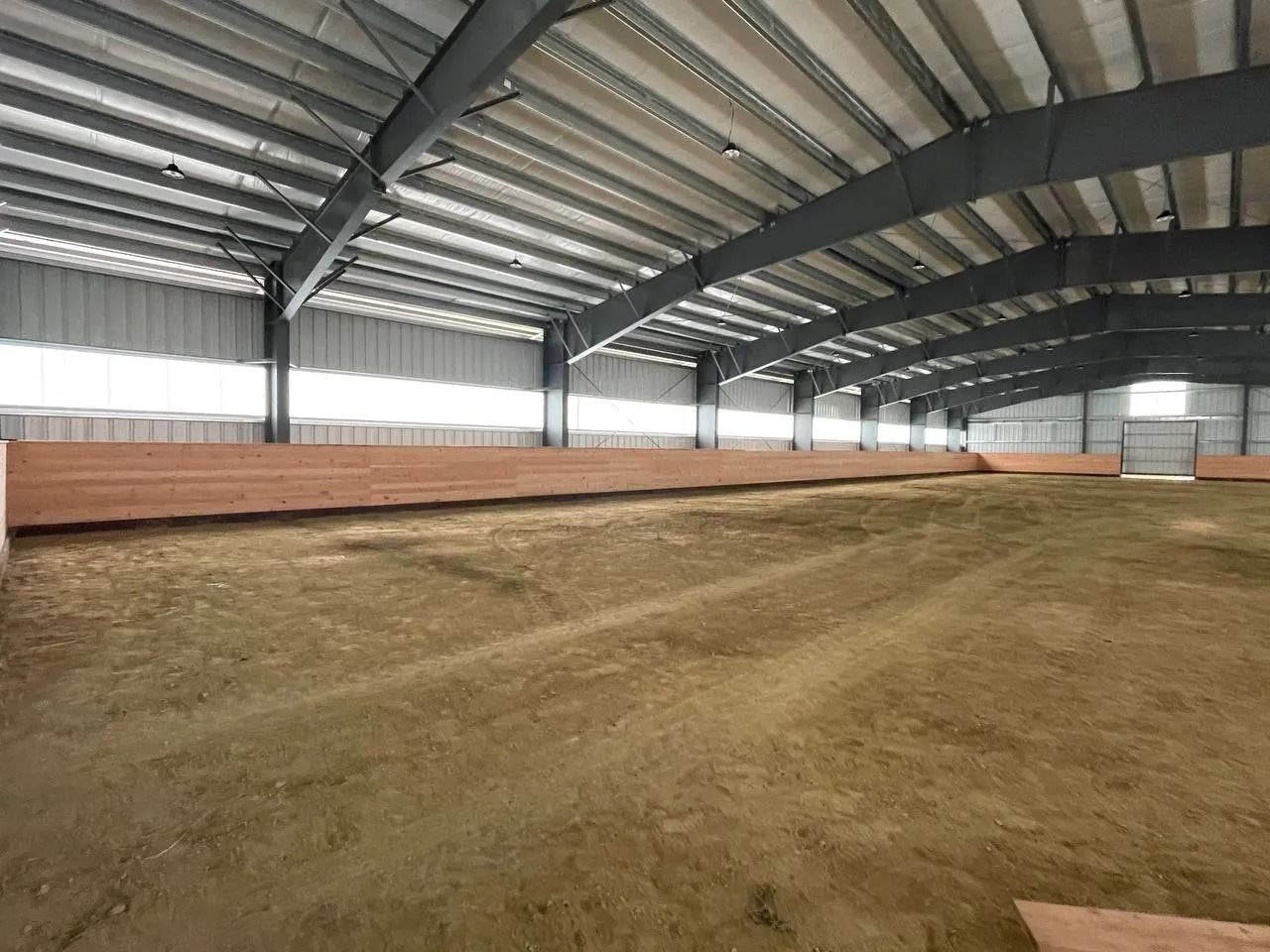 Indoor equestrian arena with tan dirt floor, wooden kickboards, and gray metal structure.