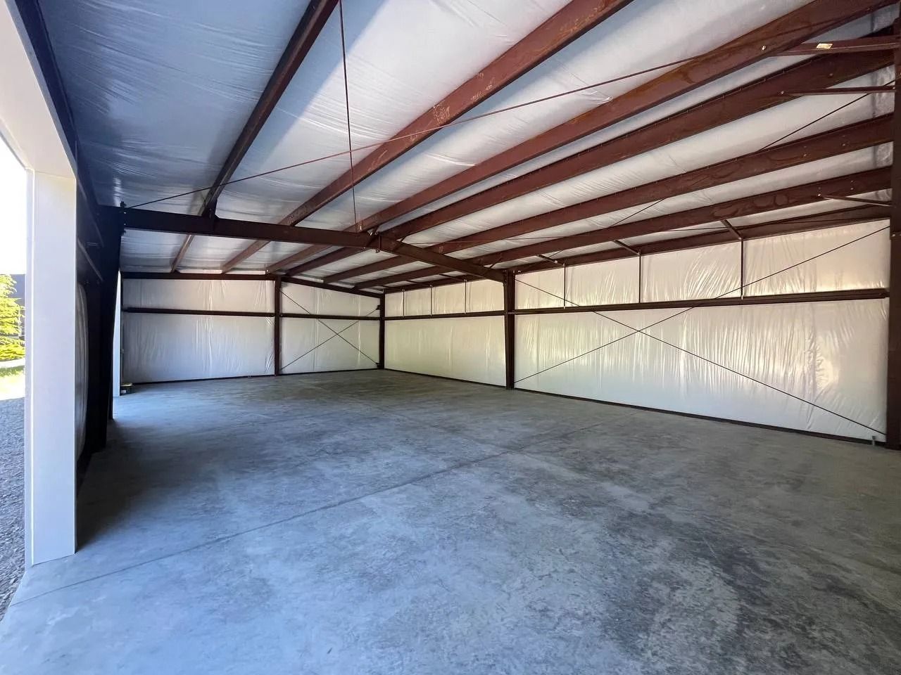Inside of an empty, concrete-floored metal building with brown beams and white insulated walls.