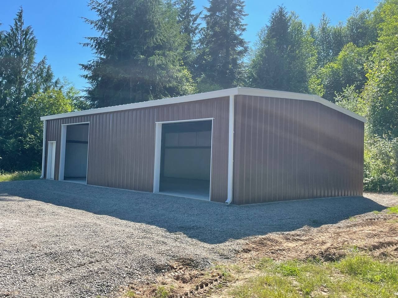Brown metal shed with two garage doors on a gravel surface, trees in background.