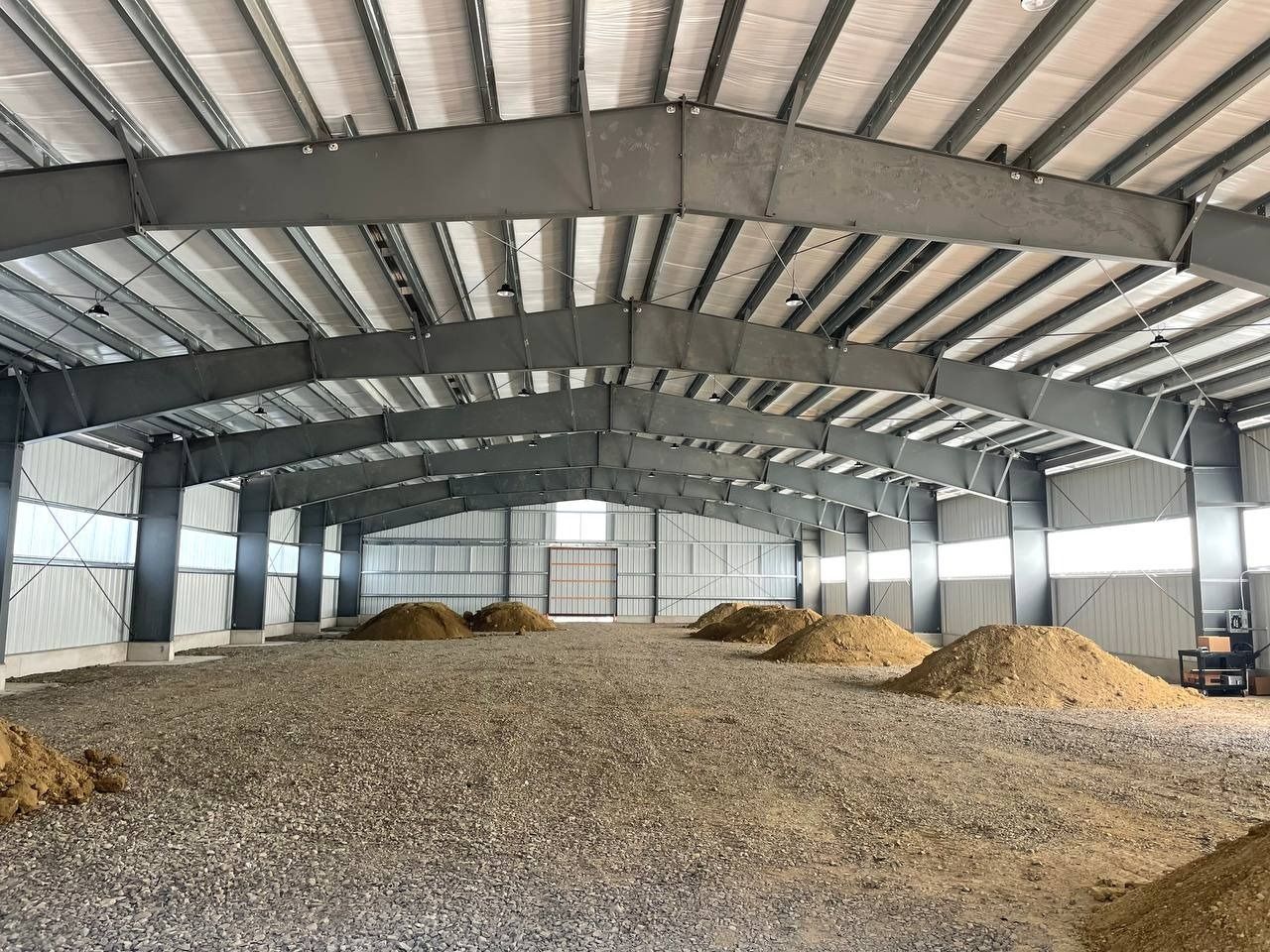 Inside of a large, empty gray metal building, dirt and gravel floor, piles of dirt.