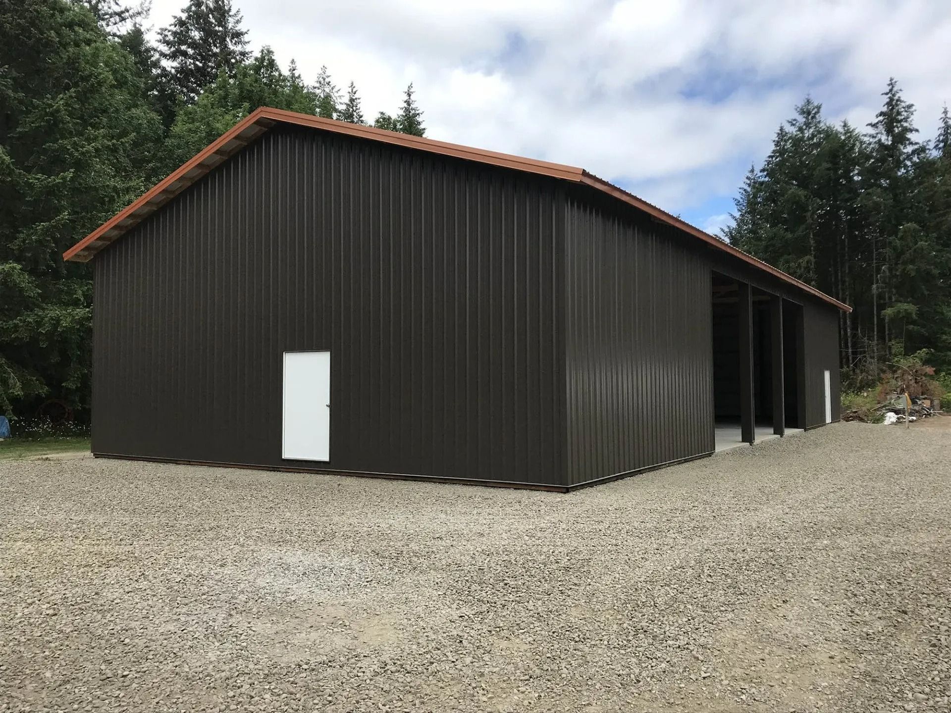 Dark brown metal building with a red roof, white door, and gravel driveway in a wooded area.