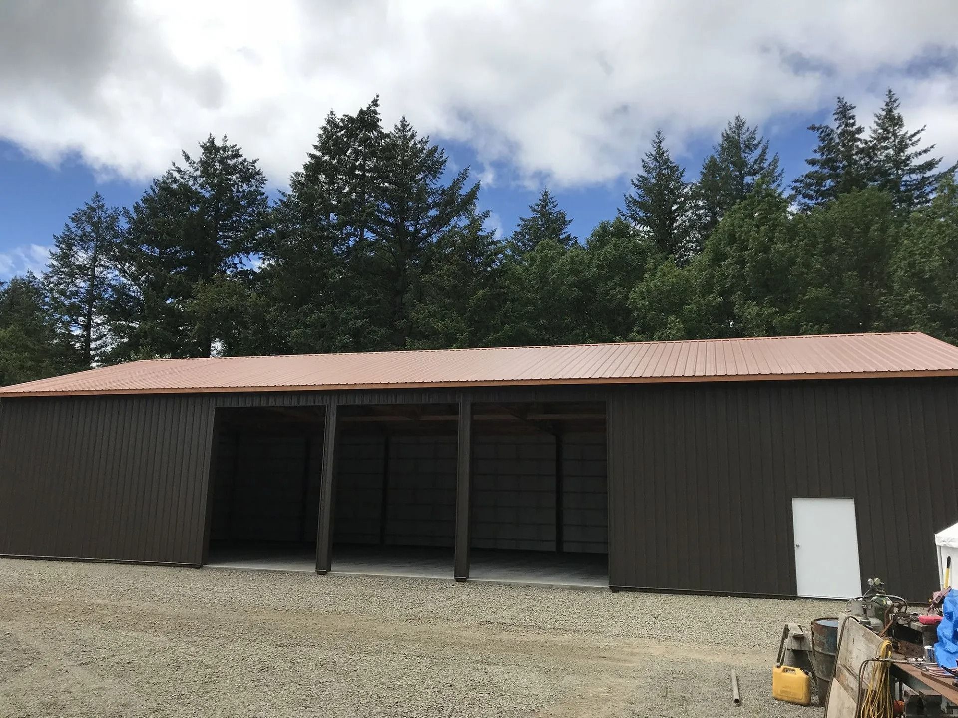 Brown metal shed with open bays, white door, gravel ground, trees, blue sky.