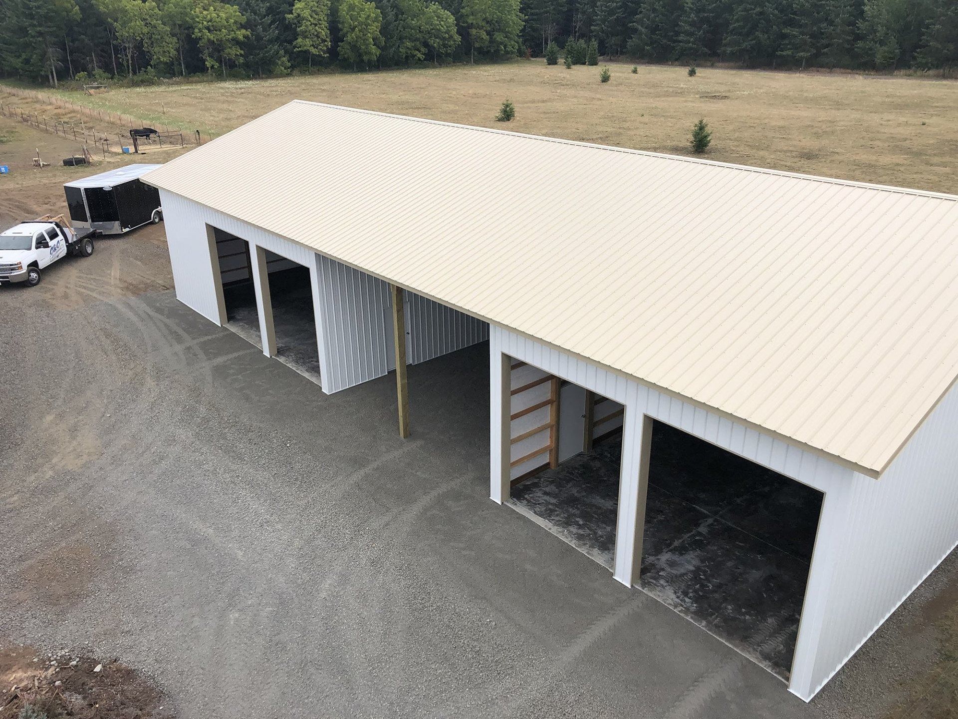 White metal building with tan roof; three open bays, gravel driveway, truck, trailer, and field.