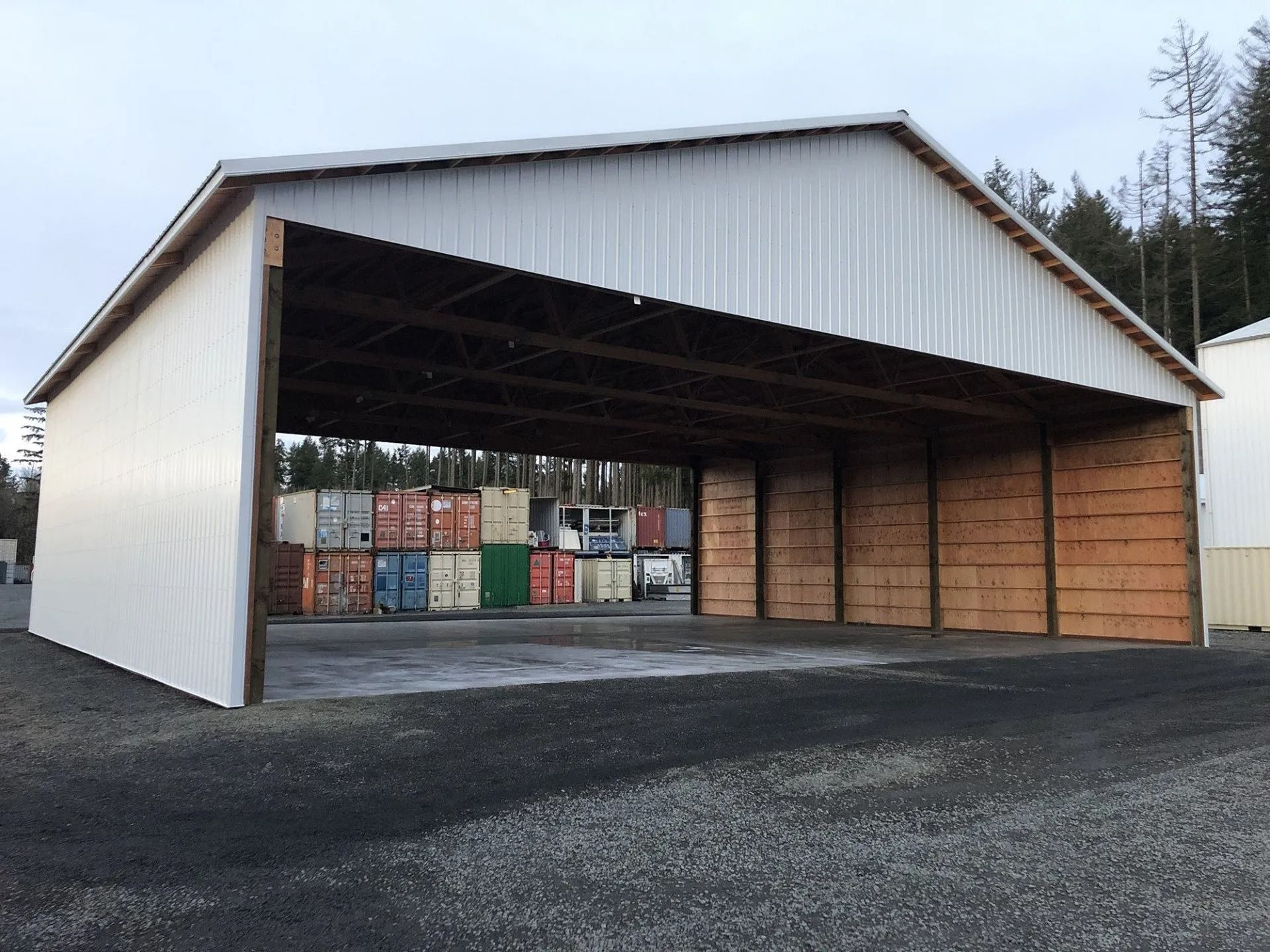 Open-sided white metal warehouse with brown wooden supports, gravel ground, and shipping containers in the background.