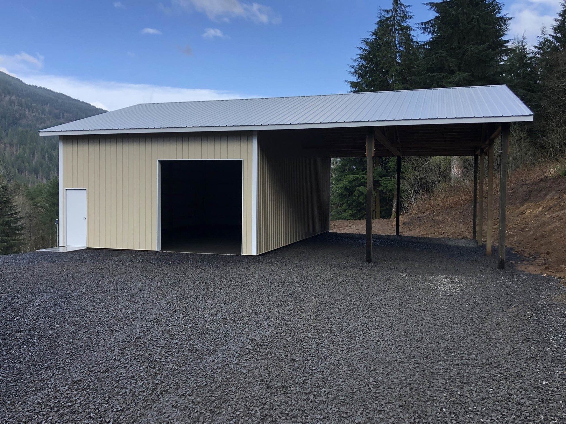 Beige metal building with a carport, gravel driveway, and trees in the background.