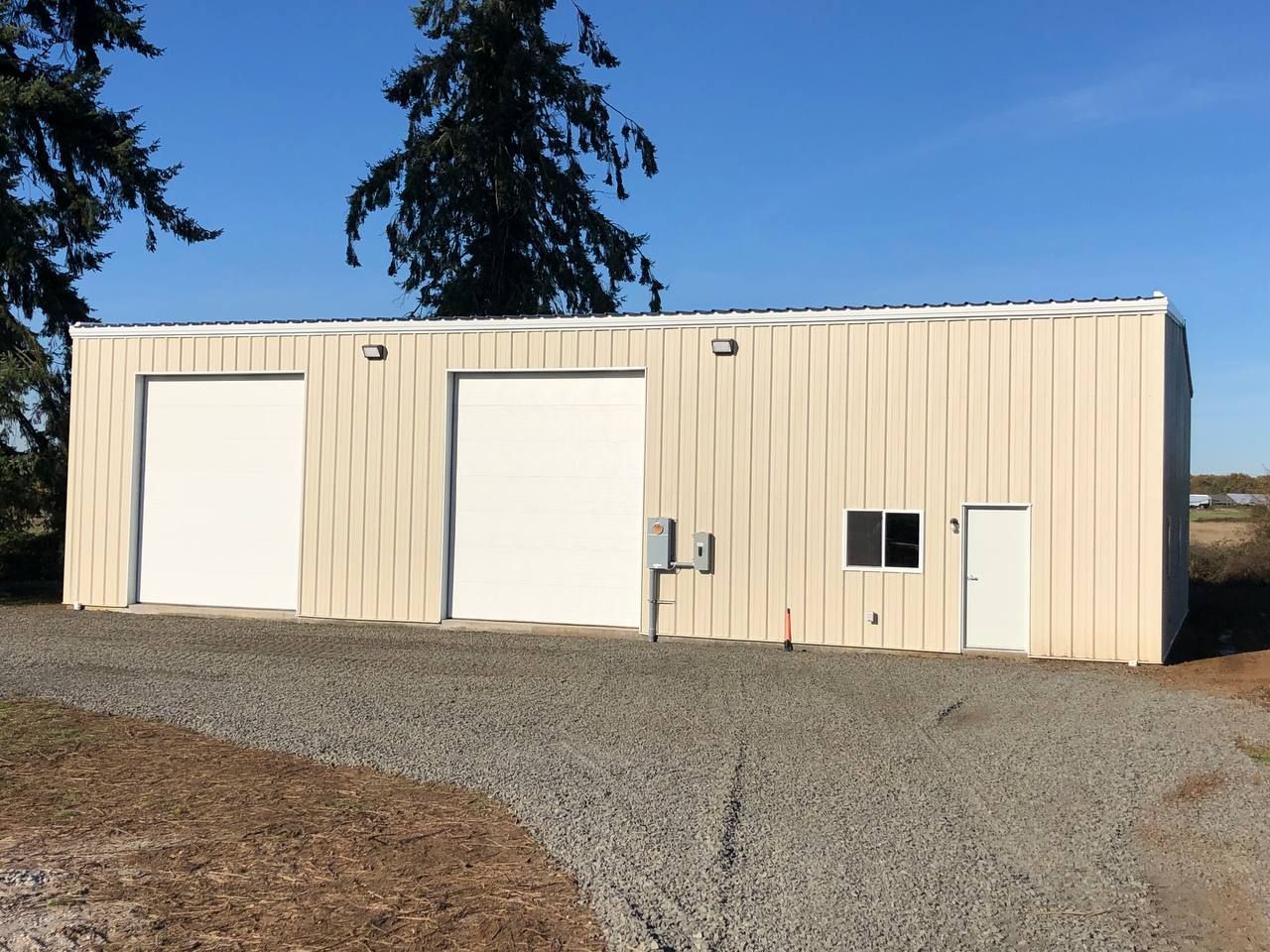 Tan metal building with three garage doors, a small window, and a single door. Gravel driveway.
