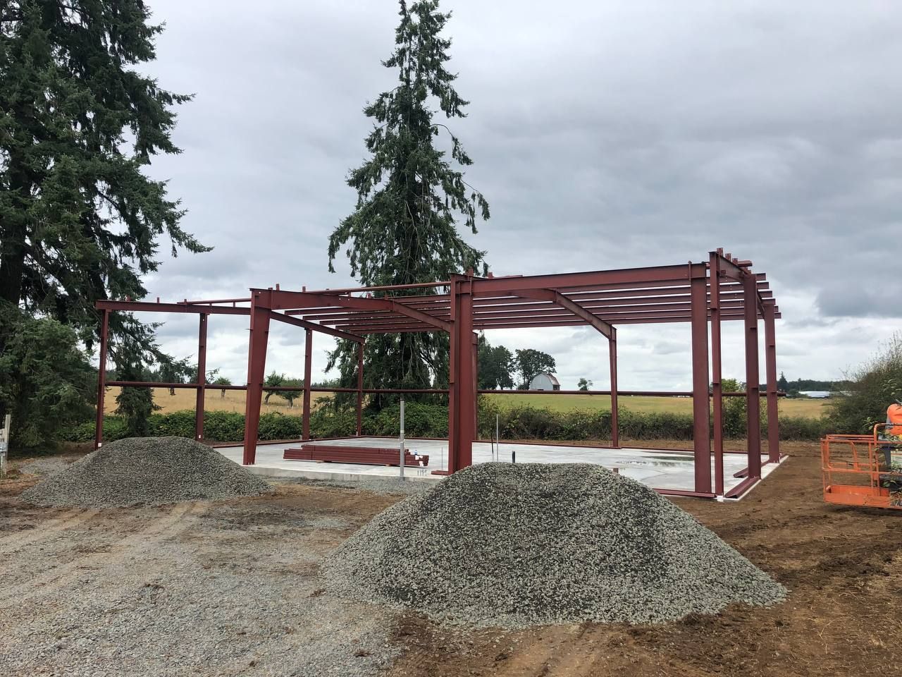 Steel frame of a building under construction, red-brown, set on a concrete pad with gravel piles in the foreground, outdoors, cloudy sky.