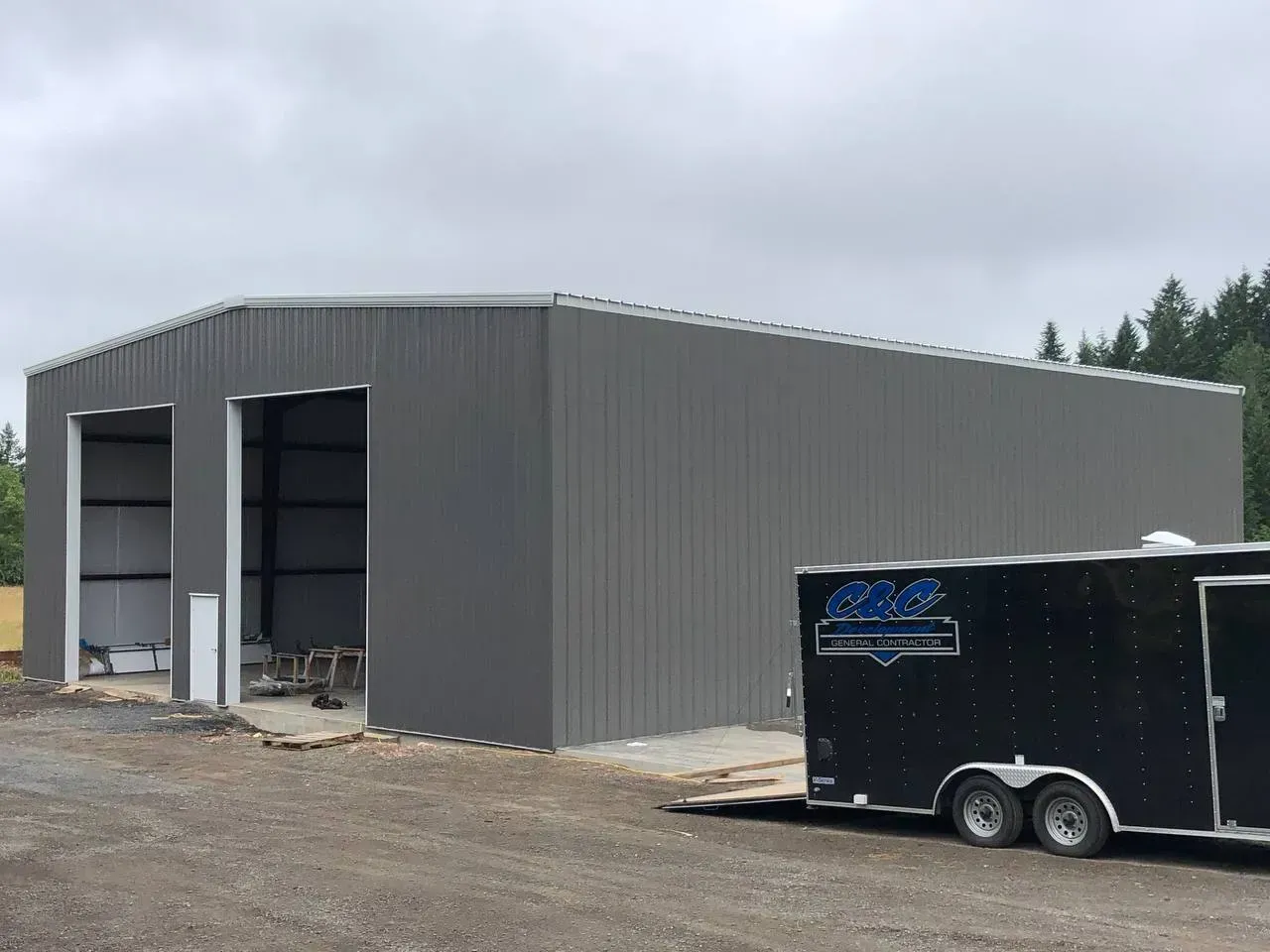 Gray metal building with two open garage doors; a black trailer is parked in front.