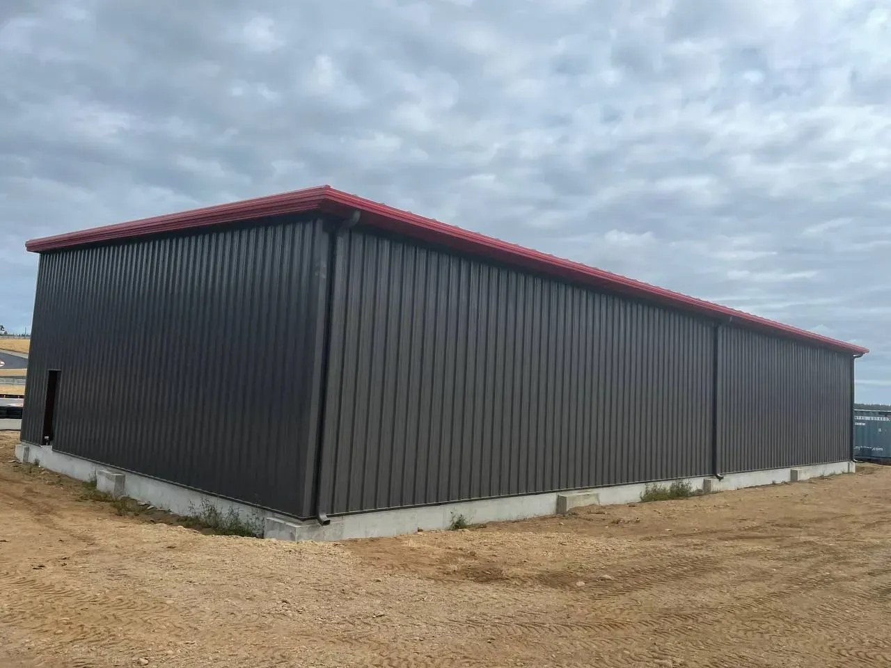 Dark grey metal building with a red roof, sitting on a concrete foundation, under a cloudy sky.
