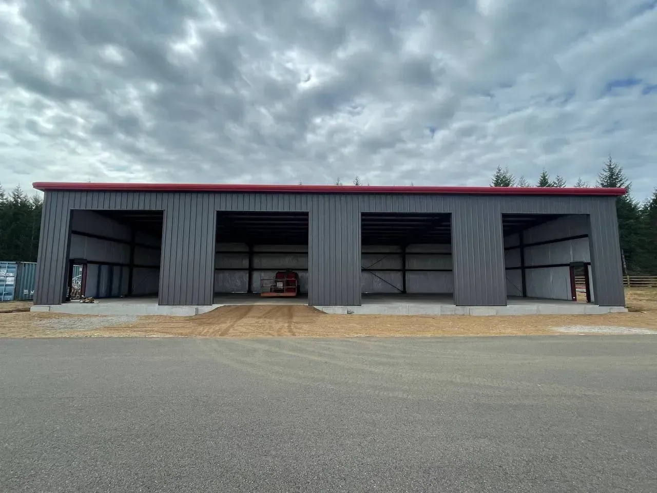 A large, gray metal shed with four open bays, a red roof, and a gravel driveway under a cloudy sky.