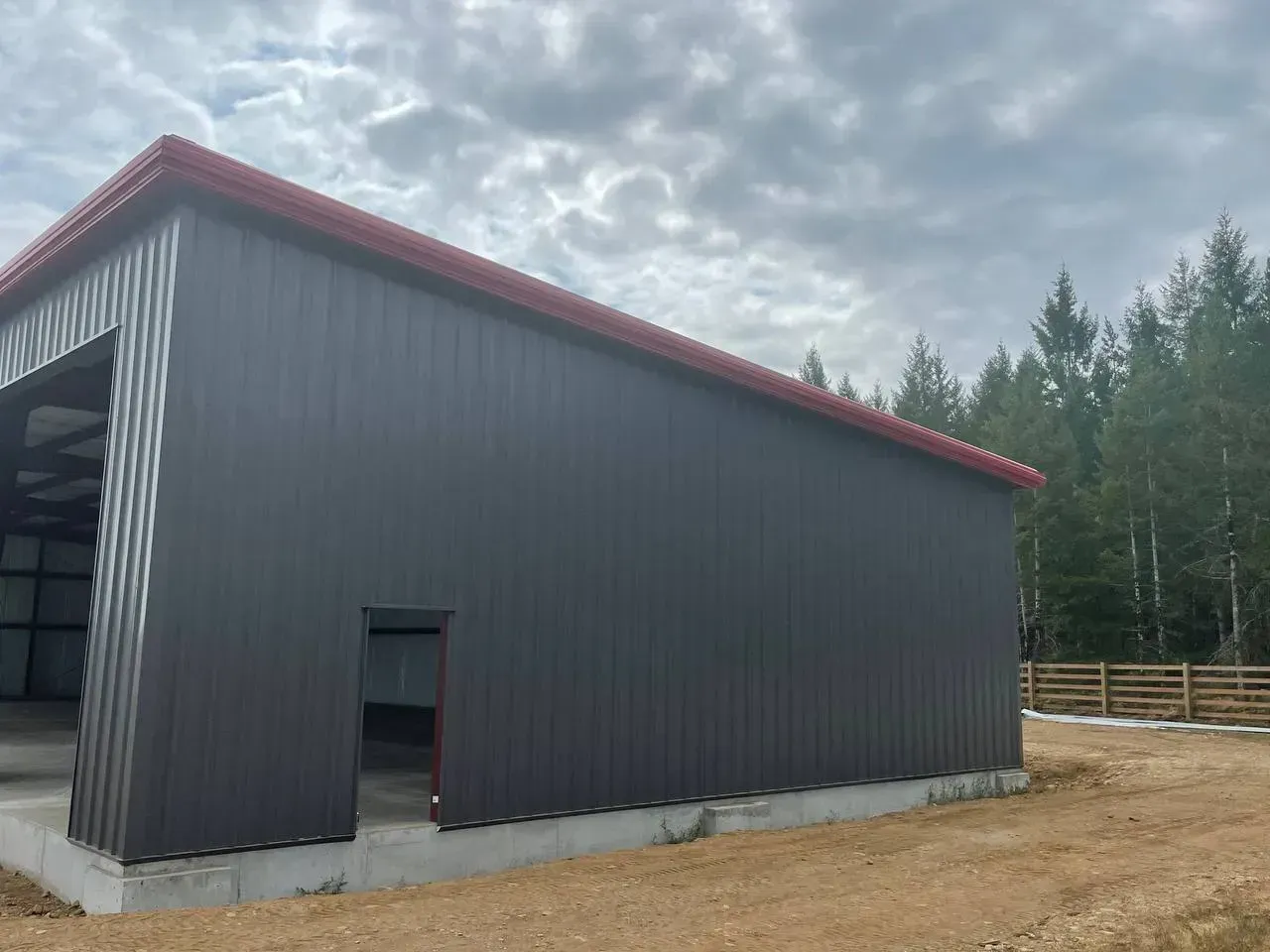 Dark gray metal building with a red roof, small door, and open bay. Forest in background.