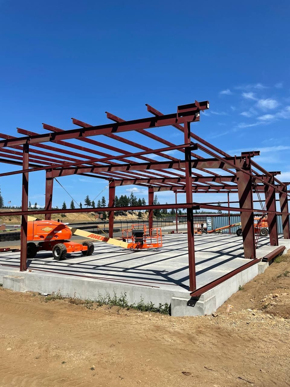 Steel frame of a building under construction on a concrete foundation, orange lifts in foreground, blue sky.