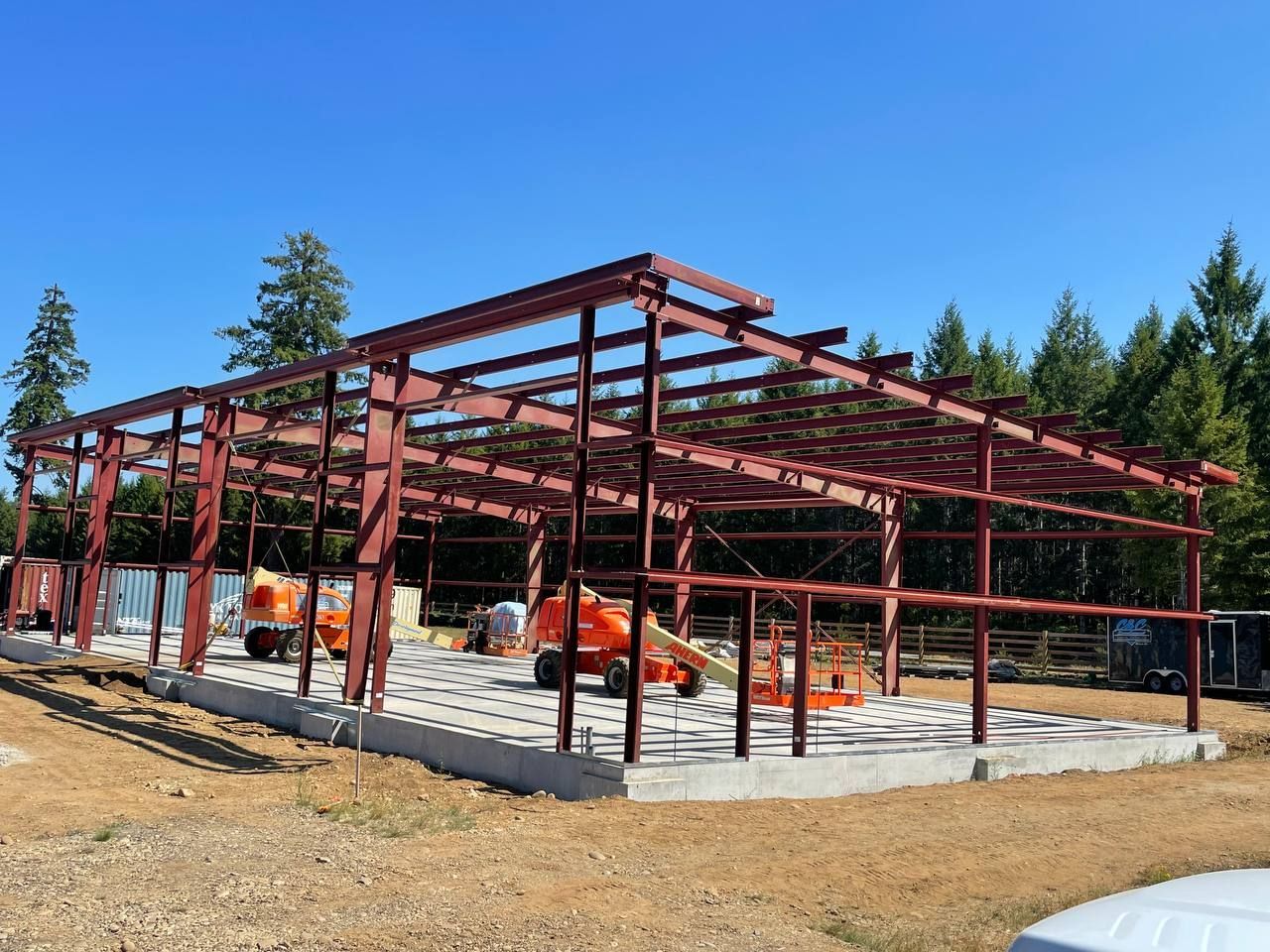 Red steel frame of a building under construction on a concrete foundation, orange lifts nearby, with trees in the background.