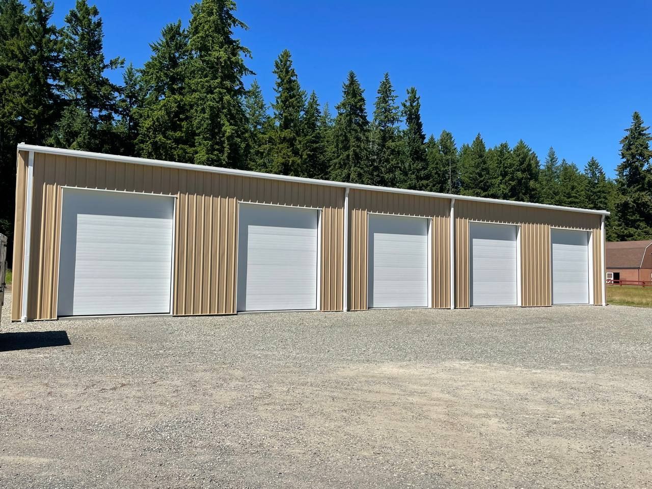Row of beige storage units with white garage doors, set against a gravel driveway and a backdrop of trees under a blue sky.