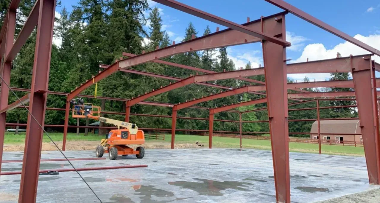 Steel frame building construction site with worker on a lift, against a green forest and sky.