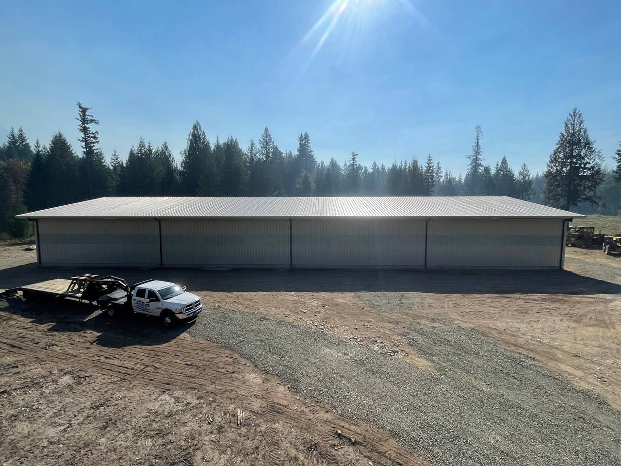 Large metal building in a gravel area with a truck and trailer, surrounded by trees under a sunny sky.
