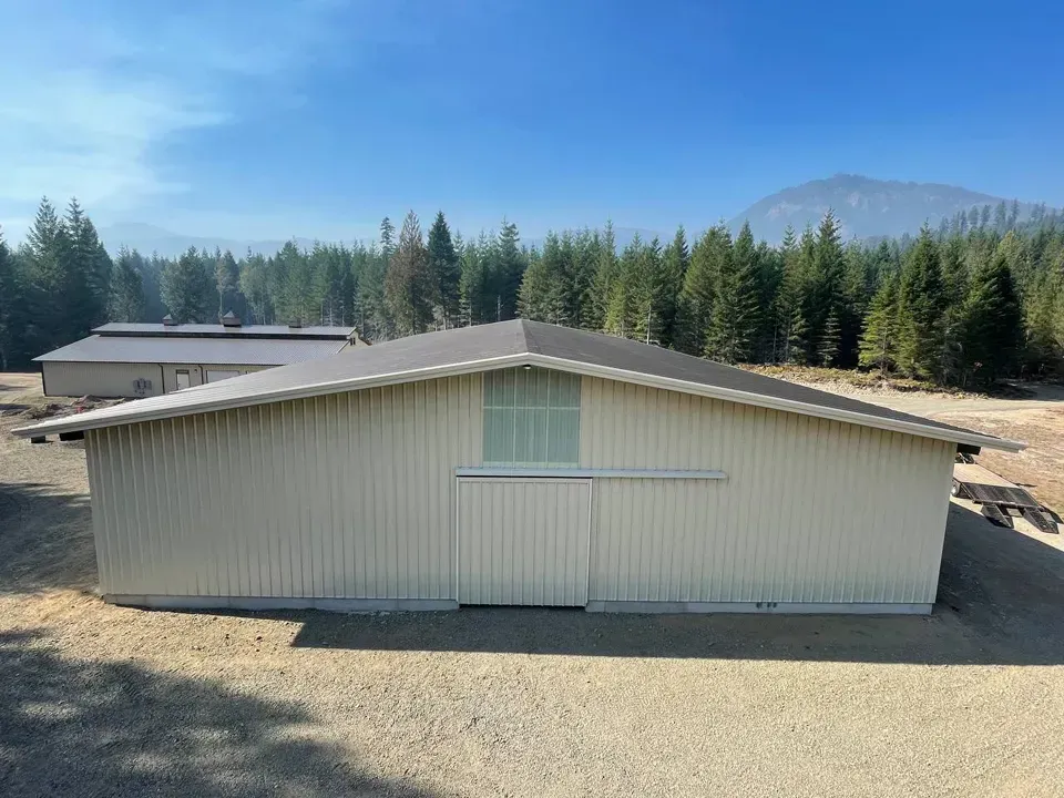 Cream-colored barn with sliding door, set against a forest and mountain backdrop under a hazy blue sky.
