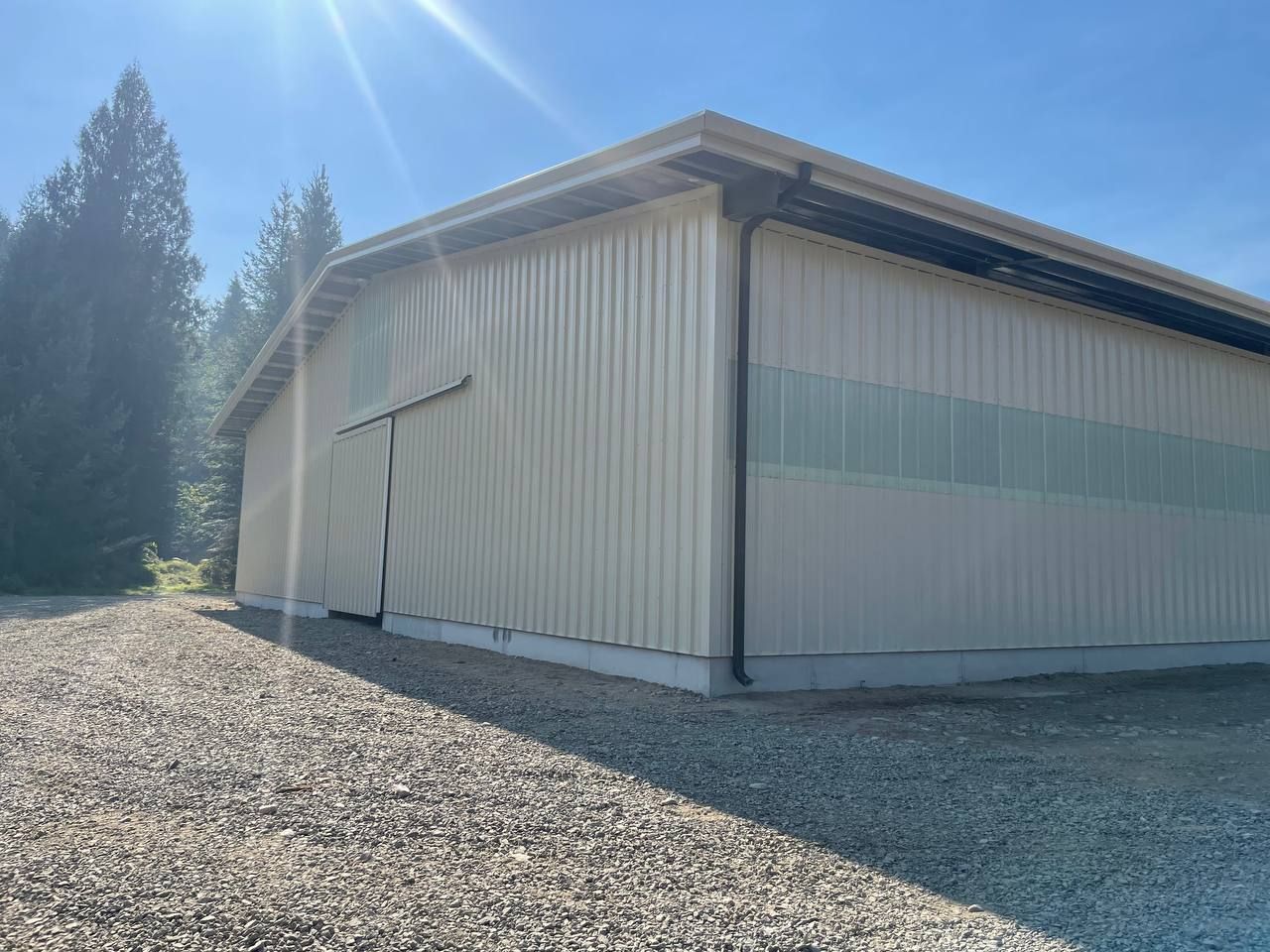 Tan metal building with green stripes, gravel ground, and trees in background.