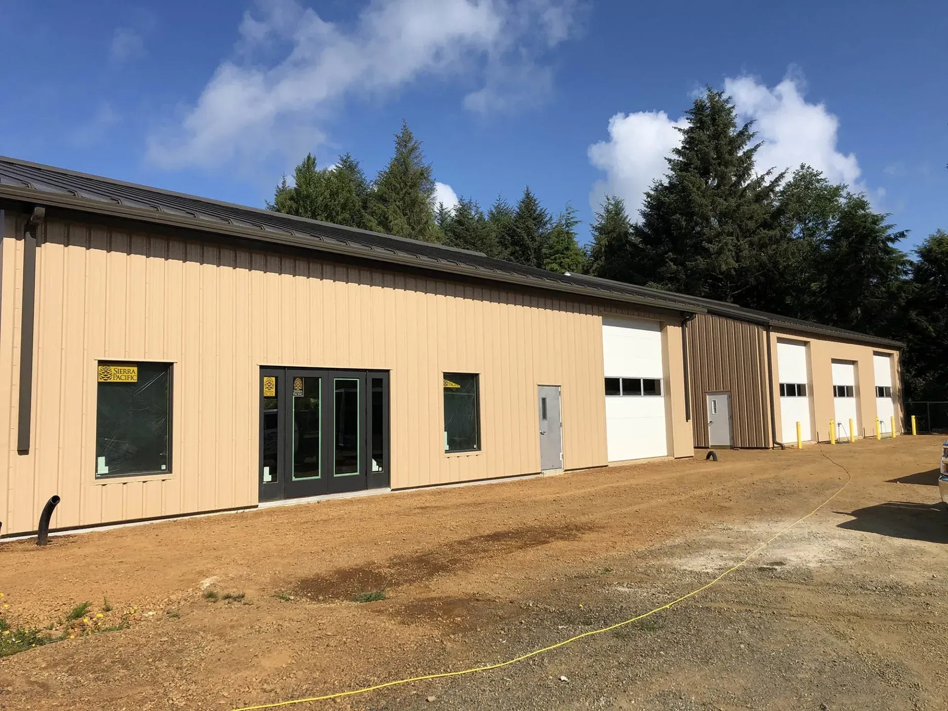 Beige industrial building with three garage doors and new construction. Gravel driveway.