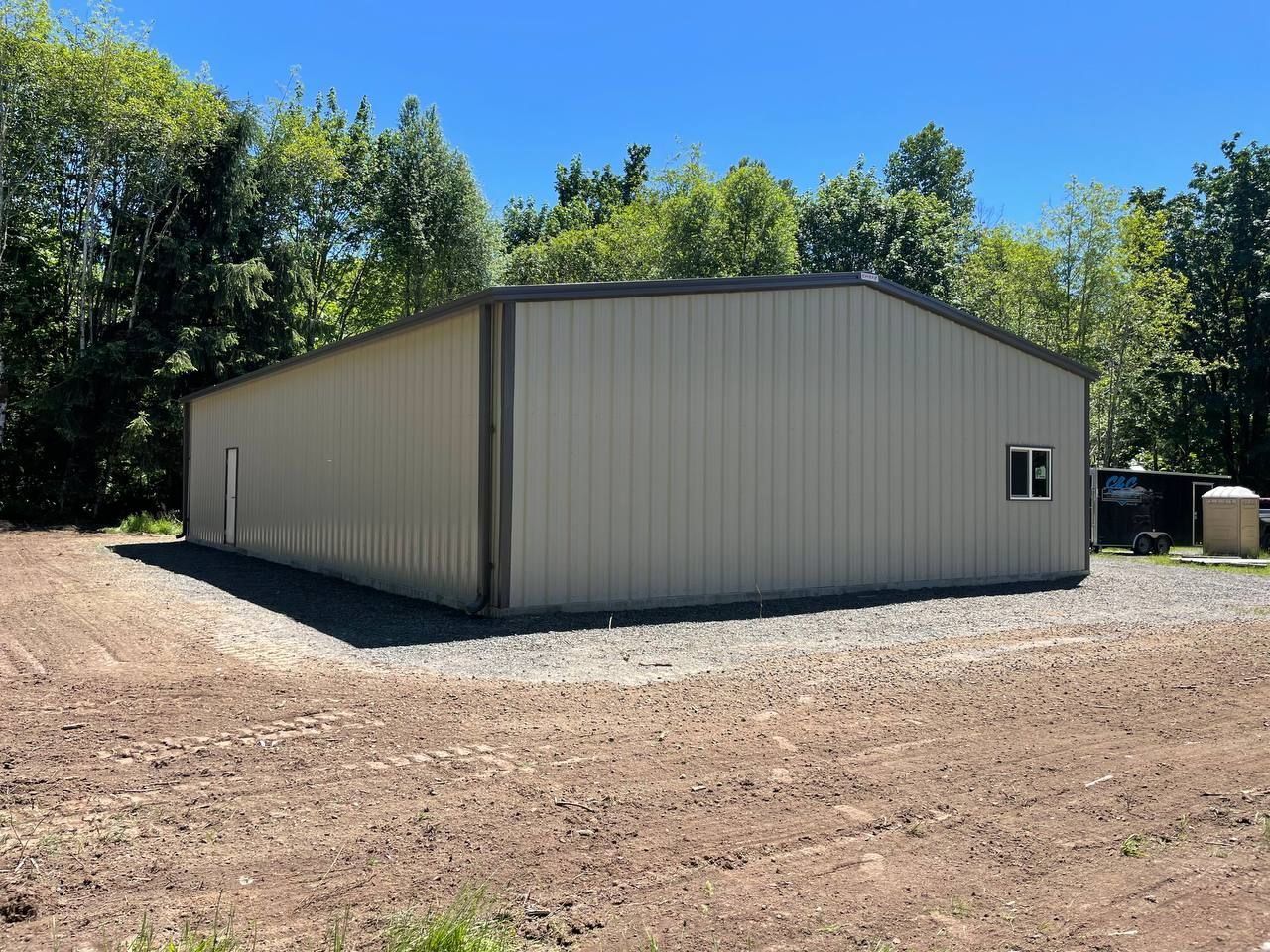 Tan metal building on gravel, small window, trees in background, sunny day.
