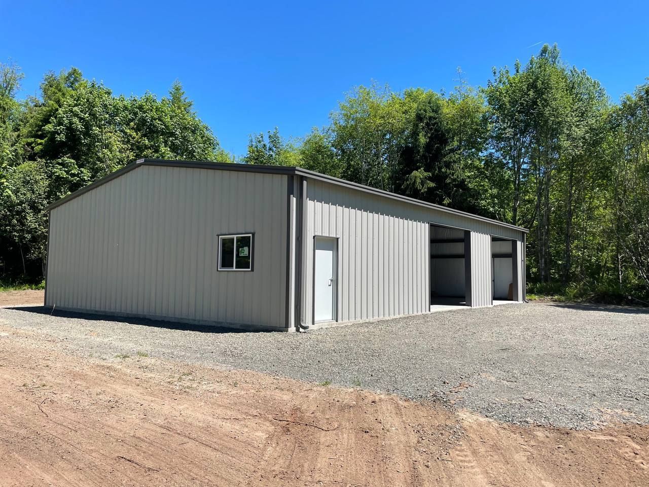 Gray metal storage building with open bay under a blue sky, surrounded by trees.