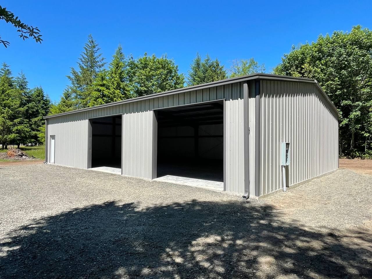 Gray metal building with two open bays, gravel ground, and trees in background.