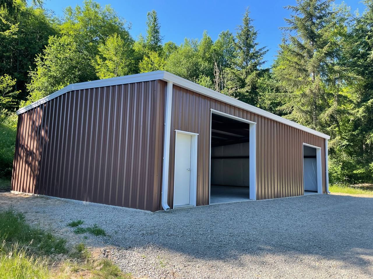 Brown metal shed with open garage door, gravel ground, and surrounding trees under a blue sky.