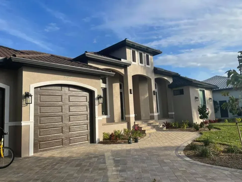 Modern beige suburban house with a garage, arched entry, and stone-paved driveway under a blue sky