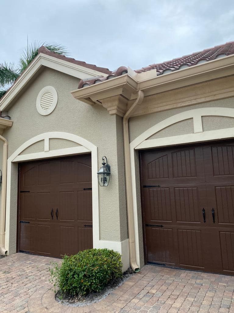 Two brown garage doors on a beige stucco house with white trim and a brick driveway