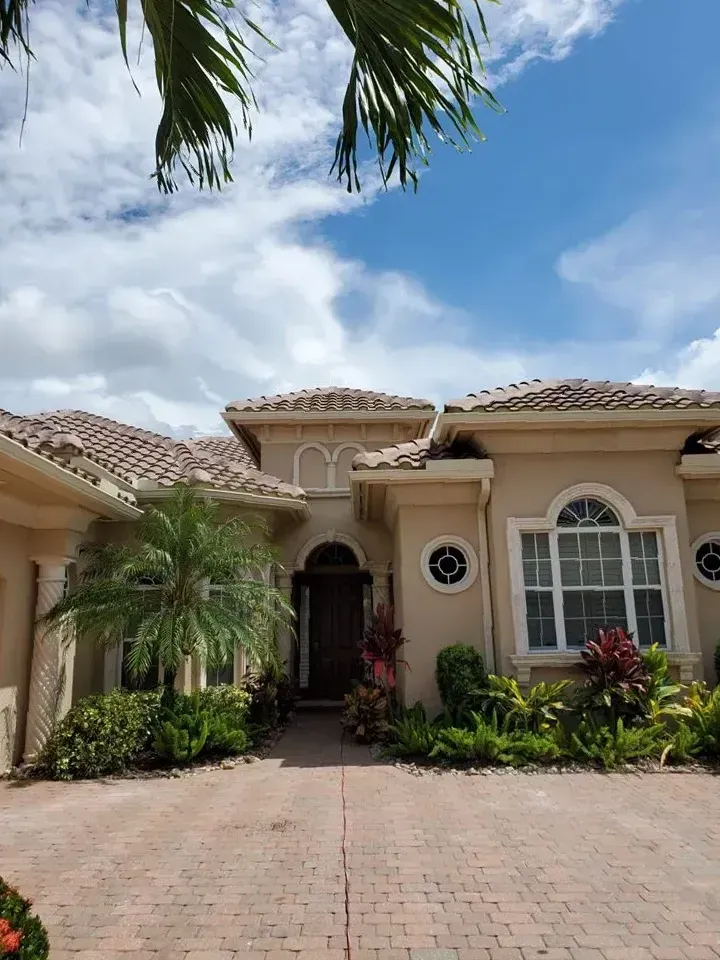 Single-story beige stucco house with a tiled roof, arched windows, and a brick driveway under a blue sky