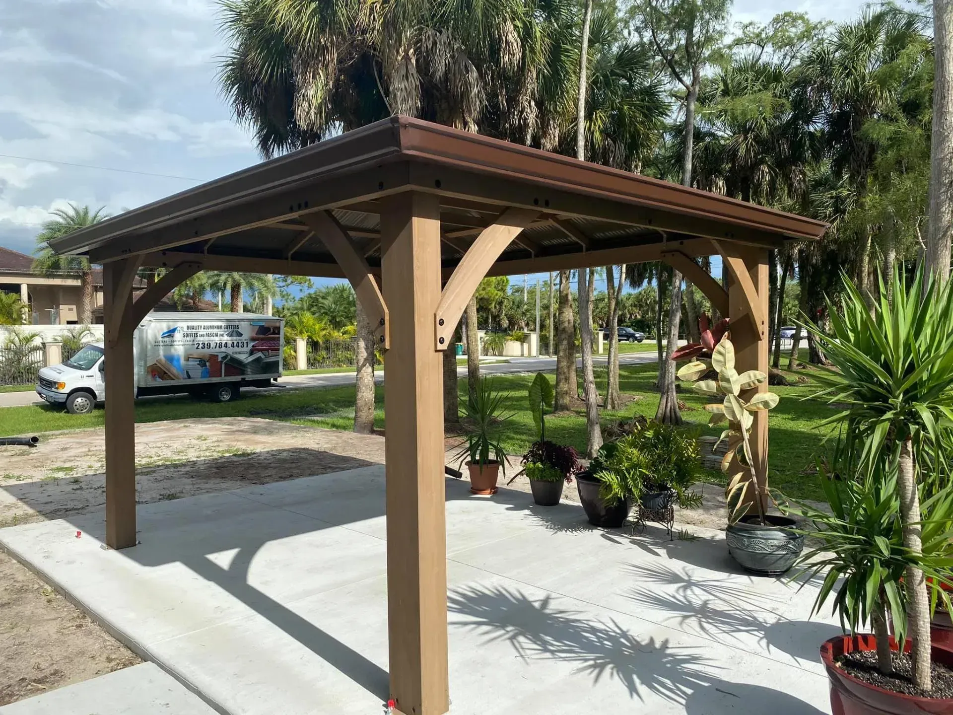 Wooden gazebo in a sunny backyard with potted plants and palm trees nearby