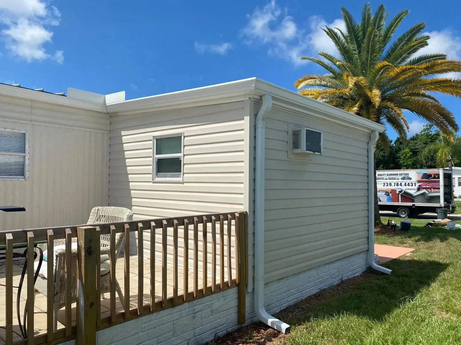 Small beige mobile home with a wooden porch fence under a blue sky and palm tree