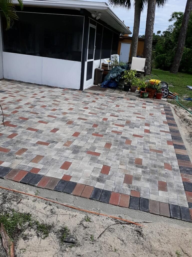 Patio with patterned pavers beside a black-and-white house, with plants and palm trees nearby