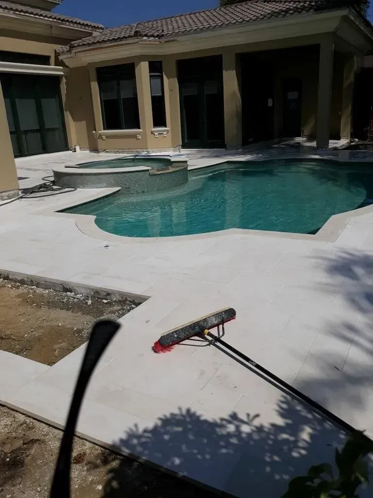 Backyard pool with a concrete patio and a pool brush lying in the foreground
