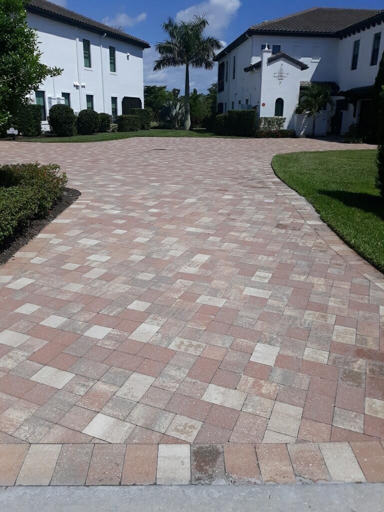 Paved driveway between white houses with red and beige brick pattern, bordered by trimmed grass and hedges