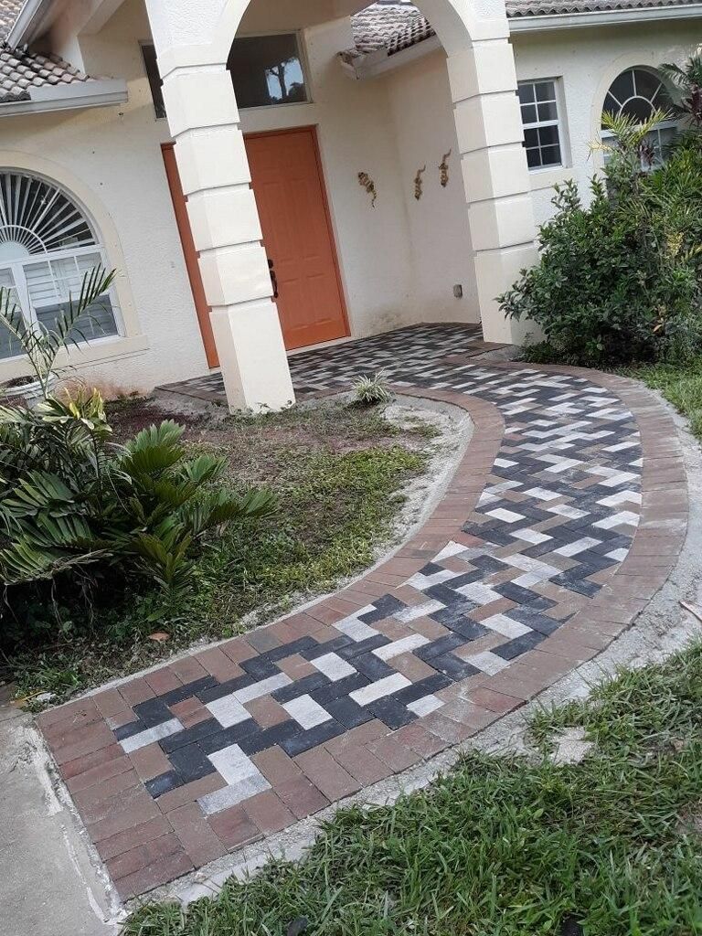 Curved brick-and-tile walkway leading to a house entrance with white columns and a peach door, bordered by grass and shrubs