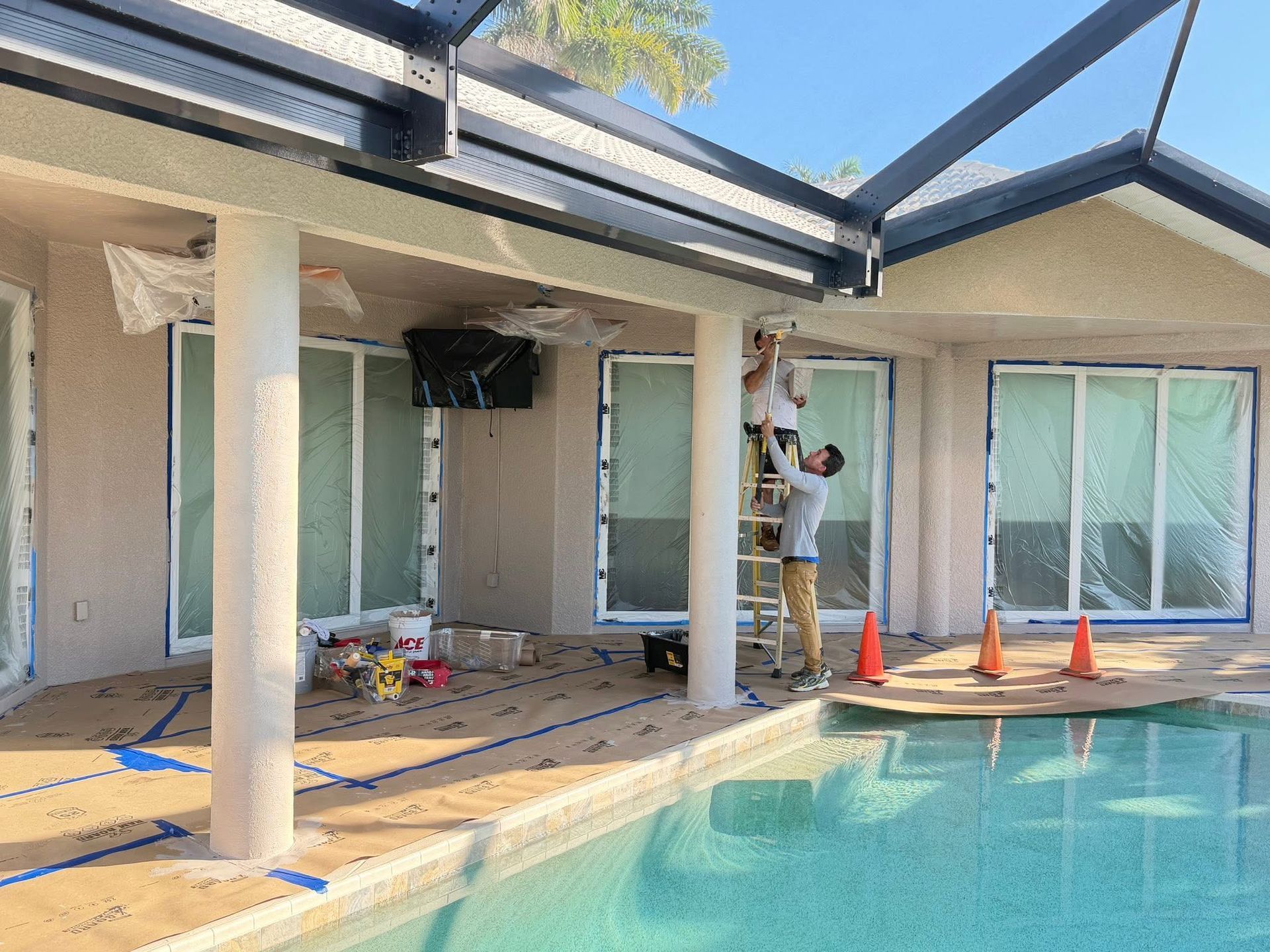 Poolside patio under construction with tools, orange cones, and a worker on a ladder under the covered area