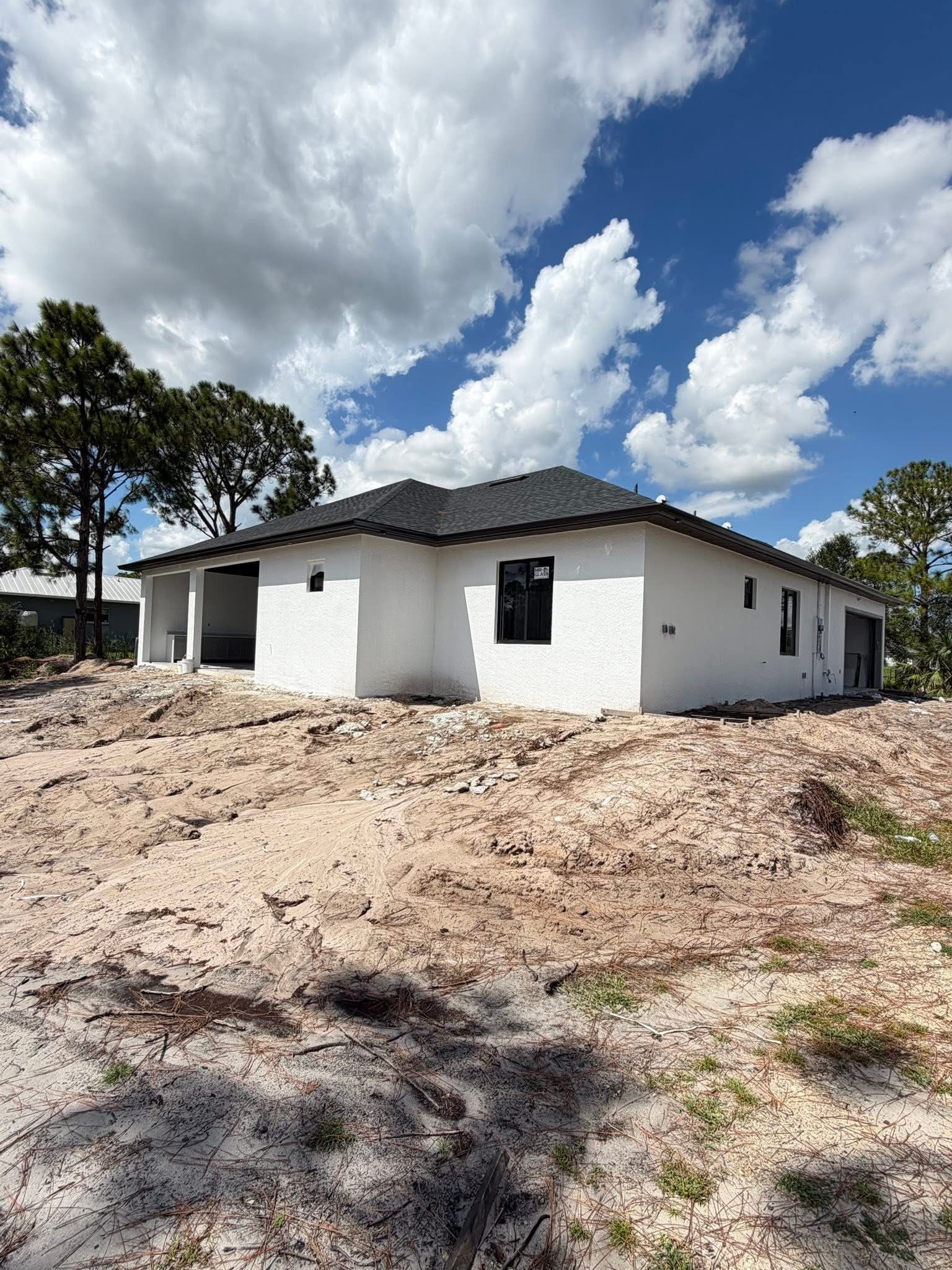 White single-story house under a cloudy blue sky, surrounded by dirt and sparse trees