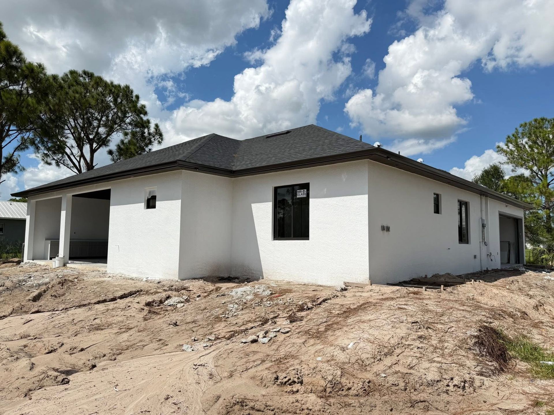 White single-story house under construction with dark roof on a dirt lot under a partly cloudy sky