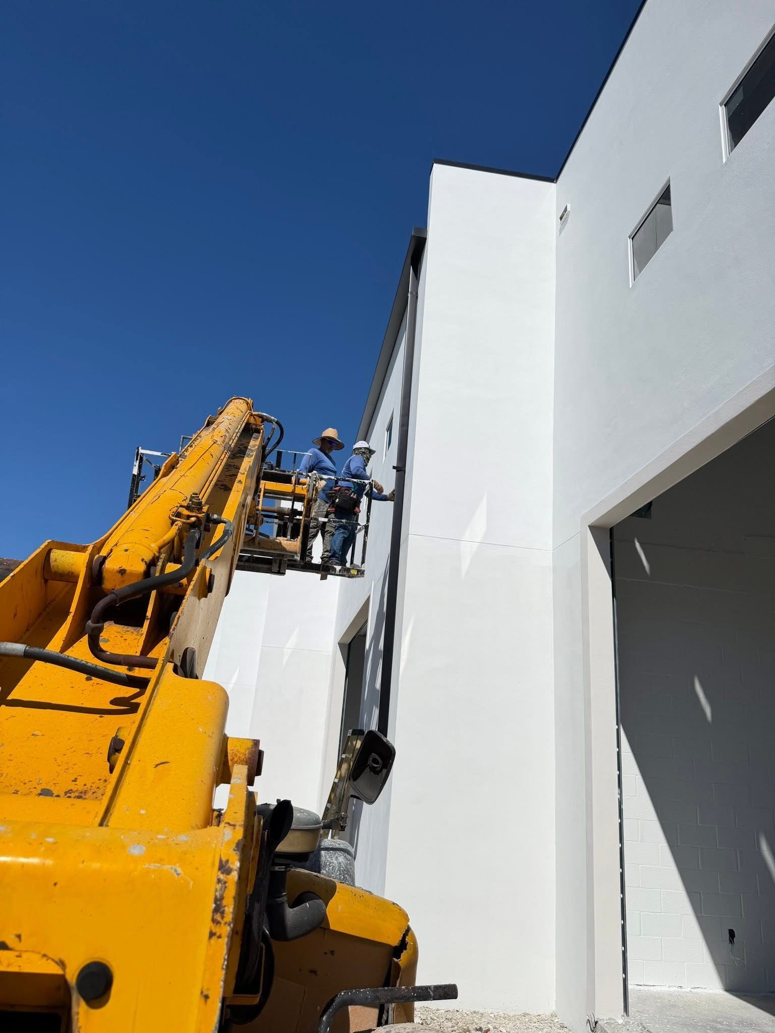 Workers on a yellow lift inspect a white building wall under a clear blue sky.