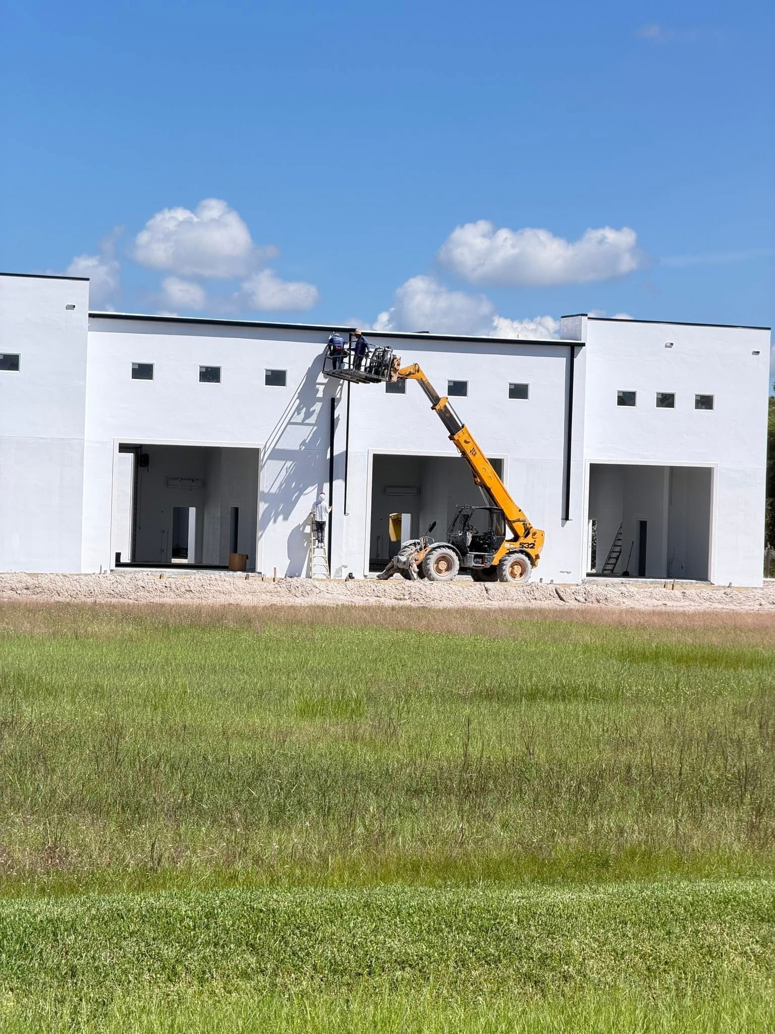 Construction worker operating a yellow boom lift outside a white industrial building under a blue sky