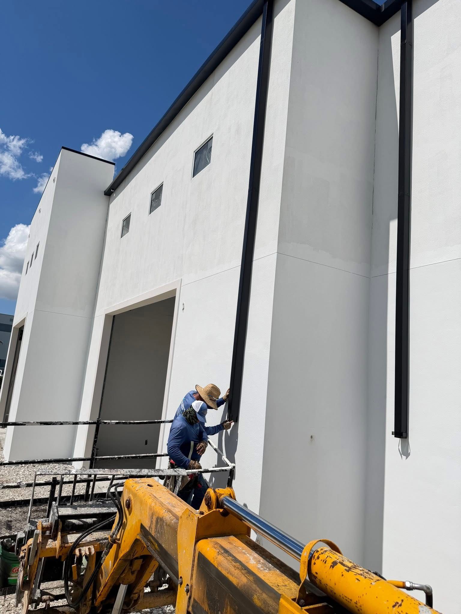 Worker on a yellow lift painting a tall white industrial building under a blue sky.