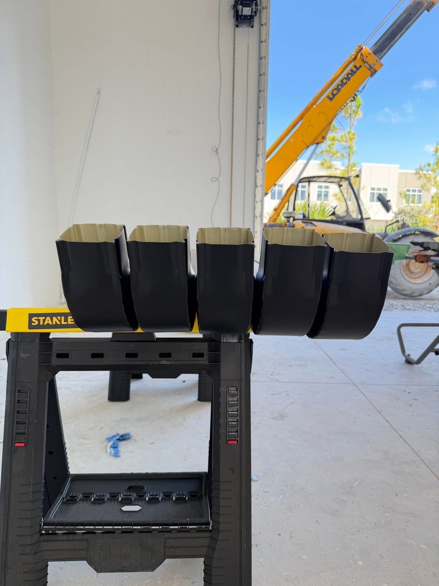 Black metal clamp fixture holding four curved black parts in a workshop with a yellow crane in the background