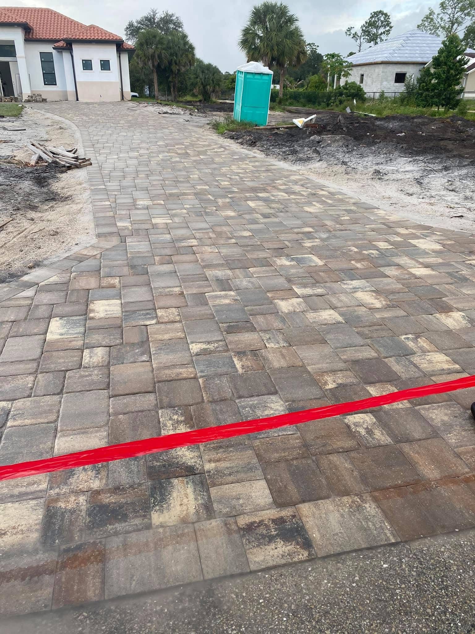 Brick-paved driveway under construction beside a house, with dirt edges and a green portable toilet in the distance