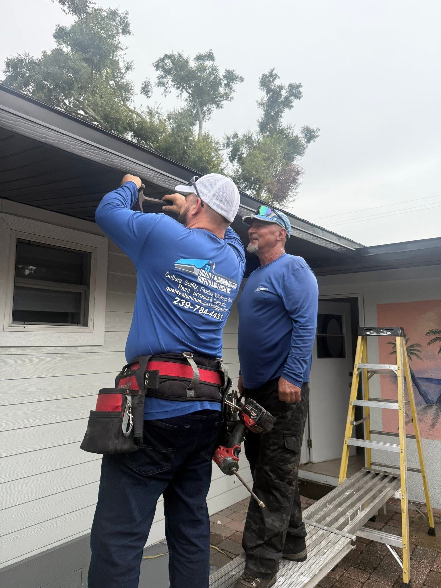 Two workers on a ladder repair roof trim beside a white house, with tools and a ladder visible.